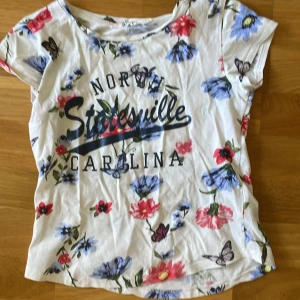 Vit blommig t-shirt från H&M - Vit t-shirt från H&M med färgglada blommor i blått och rött samt fjärilar. Tryck med texten 'North Statesville Carolina' framtill. T-shirten har korta ärmar och en klassisk rund hals. Perfekt för dig som gillar en somrig och lekfull stil.