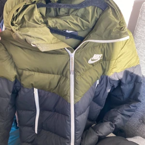 Nike pufferjacka i svart och olivgrön S - Nike pufferjacka i snygg svart och olivgrön färg med vit logga på bröstet. Jackan har huva, hel dragkedja framtill och två dragkedjeförsedda fickor. Perfekt för kalla dagar och riktigt street. Fodrad och med klassisk pufferlook.