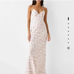 Vit långklänning med blommigt mönster - Supersöt långklänning i vit med små röda blommor och tunna axelband. Klänningen har v-ringning och figurnära passform som ger en romantisk vibe. Perfekt för varma dagar och somriga tillfällen.