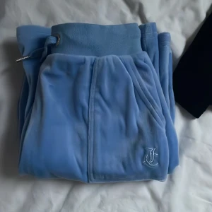Blå mjukisbyxor från Juicy Couture - Supermjuka ljusblå mjukisbyxor från Juicy Couture med broderad logga framtill och bakficka med knapp. Byxorna har bred resår i midjan och snörning för extra komfort. Perfekta för chill dagar och har den klassiska Juicy-viben. Köptes när jag var 10 så passar upp till 10 åringar. 