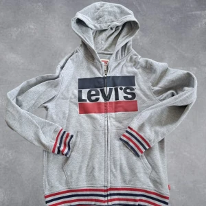 Levi's zip hoodie - Snygg grå hoodie från Levi's med stor logga framtill. Tröjan har huva, dragkedja och fickor samt ribbade muddar och nederkant med röd, blå och vit rand. Perfekt för en chill och sportig look.