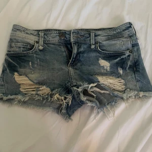 Blå slitna jeansshorts från Bershka - Riktigt snygga blå jeansshorts från Bershka med slitna detaljer och fransig kant. Klassisk femficksmodell med låg midja och dragkedja. Perfekta för sommaren när du vill ha en avslappnad och cool look.