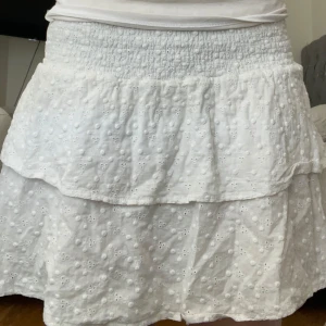 Vit volangkjol - Supersöt vit kortkjol med broderade detaljer och volanger. Kjolen har smock i midjan för en bekväm passform och är perfekt för varma dagar 