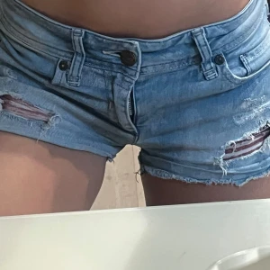Ljusblå slitna jeansshorts - Säljer ett par ljusblå jeansshorts med slitna detaljer och råa kanter. Shortsen har låg midja. Perfekta för sommaren och ger en avslappnad vibe.