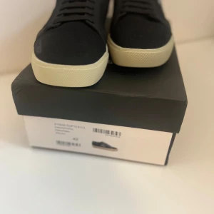 Svarta Saint Laurent sneakers strl 42 - Snygga svarta sneakers från Saint Laurent med vit logga broderad på sidan och gulddetaljer på plösen. Skorna har en klassisk låg profil, svarta snören och vit platt sula. Tillverkade i canvas och skinn för en stilren och lyxig känsla.