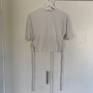 Vit croppad topp med knyt från Nelly - Säljer en vit croppad topp från Nelly i bomull med korta ärmar och snygga knytband i sidorna som kan knytas i ryggen. Perfekt för dig som gillar trendiga detaljer och en enkel stil. Den är aldrig använd💗