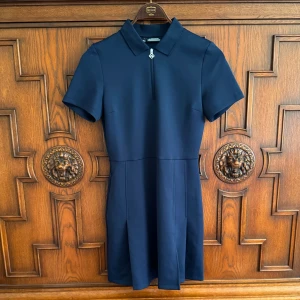 Marinblå sportig klänning J.Lindeberg - Sportig marinblå kortklänning från J.Lindeberg med krage, kort ärm och halv dragkedja framtill. Klänningen har en stilren design med diskreta sömmar och medföljande matchande shorts. Perfekt för en aktiv och snygg look.