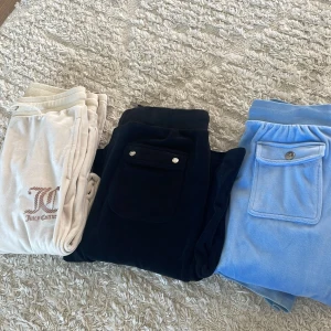 Juicy Couture velourbyxor i 3 färger - Tre par klassiska Juicy Couture mjukisbyxor i velour – ett par ljusblå, ett par svarta och ett par cremevita. Alla har snörning i midjan, bakfickor med knapp och bootcut-snitt. Perfekta för en chill och trendig look. Dom svarta är storleken 12/13 och dom beige är också det och dom blå är 10/11 