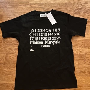Maison Margiela svart t-shirt med tryck - Svart t-shirt från Maison Margiela med ikoniskt vitt tryck framtill med siffror och logga. Klassisk passform, rund hals och korta ärmar. Tillverkad i mjuk bomull som känns skön mot huden. Perfekt för dig som gillar stilrena och trendiga plagg med edge.