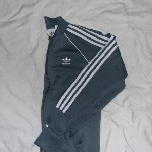 Adidas svart track jacket med ränder - Snygg askgrön Adidas track kofta med klassiska vita ränder längs ärmarna och broderad logga på bröstet. Koftan har dragkedja framtill, ribbade muddar och hög krage. Perfekt för en sportig och avslappnad stil.