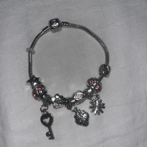 Silverfärgat berlockarmband från Pandora - Charmigt armband i silverfärg från Pandora med flera berlocker, bland annat en nyckel, jordgubbe, blomma och stjärna. Armbandet har detaljer i rött och rosa samt glittriga pärlor. Perfekt accessoar för att ge din stil en personlig touch.