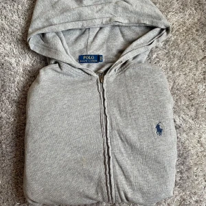 Grå hoodie från Polo Ralph Lauren - Snygg grå hoodie från Polo Ralph Lauren i 100% bomull. Klassisk modell med dragkedja, huva och mörkblå broderad logga på bröstet. Perfekt för en chill och stilren look. Passar dig som gillar enkel och tidlös streetstyle.