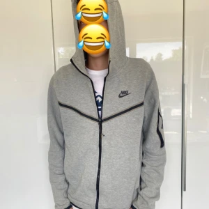 Grå zip-hoodie från Nike - Grå hoodie från Nike med hel dragkedja, svarta detaljer och ficka med dragkedja på ärmen. Klassisk logga på bröstet och huva. Perfekt för chill eller när du är på språng.