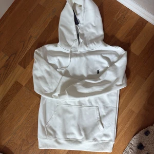 Vit hoodie från Polo Ralph Lauren - Säljer en stilren vit hoodie från Polo Ralph Lauren med klassisk huva och snörning. Tröjan har en broderad logga på bröstet och en stor magficka. Perfekt för dig som gillar enkel och clean streetstyle.
