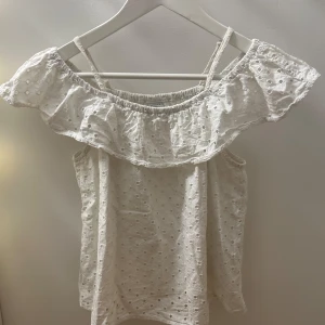 Vit offshoulder blus från Lindex - Söt vit blus från Lindex med offshoulder och smala axelband. Blusen har broderade hålmönster och en bred volang runt axlarna. 