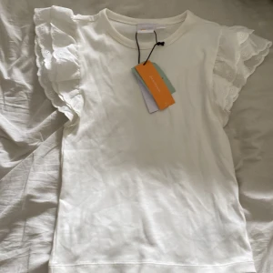 Vit volangtopp från Gina Tricot - Supersöt vit topp från Gina Tricot med breda volangärmar i broderad bomull. Toppen är ribbad i tyget och har en rund halsringning. Perfekt för dig som vill ha en feminin och trendig look med extra detaljer på axlarna.helt ny jag köpte den när jag var i Göteborg här om Dan men märkte att den var för liten.