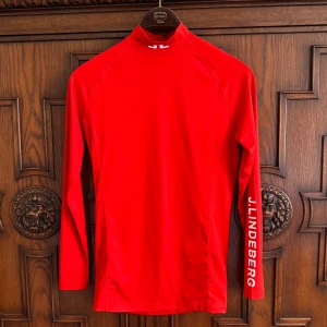 Soft kompression träningströja J.Lindeberg - Snygg röd långärmad kompression träningströja från J.Lindeberg med vit logga på ärmen och halsen. Tröjan har en tight passform och är gjord i mjukt, stretchigt syntetmaterial som andas. Perfekt för dig som vill ha en sportig look och gillar att sticka ut.