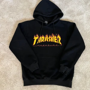 Svart Thrasher hoodie - Svart hoodie från Thrasher med ikoniskt flames-tryck i gult och rött på bröstet. Klassisk känguruficka, huva med snörning och mjukt material. Perfekt för dig som gillar streetwear och skate-stil. Saknar tyvärr ena metallbiten på en av snörningen men inget som märks.