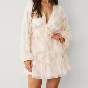 Blommig kortklänning med volang - Superfin kortklänning med blommigt mönster i ljusa pastellfärger. Klänningen har v-ringning, långa ärmar och volangdetaljer i kjolen. Perfekt för sommarens alla tillfällen och ger en romantisk vibe.