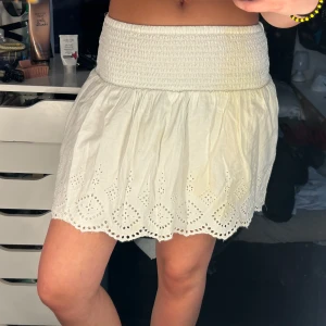 Vit kortkjol med broderade detaljer - Supersöt vit kortkjol med bred smock i midjan och broderade hålmönster längs fållen. Kjolen är luftig och har en lätt volangig kant, perfekt för varma dagar. Tyvärr så råkade jag spilla på den, så i vissa ljus (som detta) så syns de lite. Den är från Gina Young och i stl 146/152 men passar mig som har xs-s🥰.