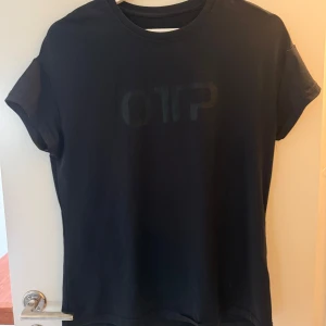 Svart t-shirt från OTP i träningsmaterial - Svart tränings t-shirt från OTP med diskret logga framtill. Klassisk rund halsringning och korta ärmar. Tillverkad i mjukt träningsmaterial som känns skön mot huden. Perfekt för en enkel stil.
