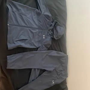 Blå vindjacka från Under Armour S - Snygg blå dress från Under Armour med luva och dragkedja. Jackan har logga på bröstet och stor Under Armour-tryck på ryggen. Tillverkad i polyester, perfekt för träning eller när du vill ha en sportig look. Långärmad och med praktiska fickor framtill.
