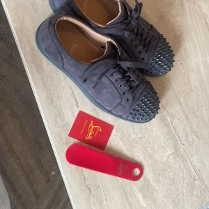 Christian Louboutin sneakers med nitar - Mörkgrå sneakers i mocka från Christian Louboutin med svarta nitar på tån och klassisk röd sula. Skorna har snörning och en rund tå, vilket ger en edgy och lyxig vibe. Perfekta för dig som vill sticka ut med exklusiva detaljer. Det ändå är den svarta pricken som hände när jag råka trampa på en cigg sen annars helt nya!