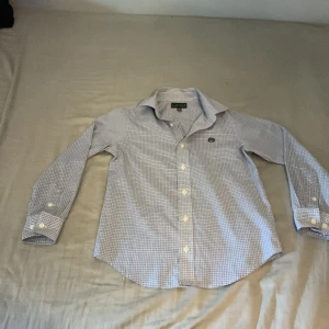 Rutig skjorta från Ralph Lauren - Snygg rutig skjorta från Ralph Lauren i blått och vitt. Skjortan har klassisk krage, långa ärmar med manschett och knappar framtill. Broderad RL-logga på bröstet. Tillverkad i mjuk bomull, perfekt för en clean och stilren look. Pris kan diskuteras