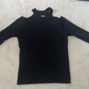 Svart ribbad polotröja med cut out - Svart ribbad polotröja med lång/trekvarts ärm och trendiga cut out-detaljer vid axlarna. Hög krage och tight passform gör den perfekt för lager på lager eller som statement-piece. Enkel att matcha med både jeans och kjol.