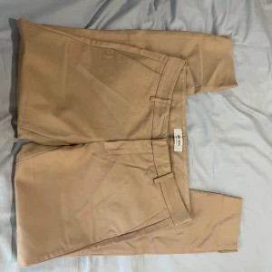 Beige kostymbyxor från Mos Mosh - Snygga beige kostymbyxor från Mos Mosh i klassisk rak modell. Byxorna har hällor för bälte och diskreta fickor framtill. Perfekta för en clean och stilren look, matcha med skjorta eller t-shirt för en chill vibe.