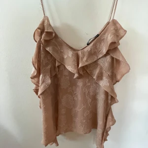 Beige volangtop från Zara - Supersöt beige linnetopp från Zara med tunna axelband och volangdetaljer längs ringningen och sidorna. Toppen har ett subtilt blommönster i tyget som ger en romantisk vibe. Perfekt för varma dagar när du vill känna dig extra fin.