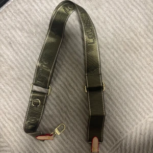 LV axelrem grön - Axelrem från Louis Vuitton i grön färg med justerbar längd och guldfärgade metalldetaljer. Remmen har präglad logga och röda detaljer vid fästena. 