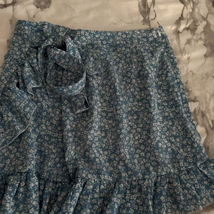 Blå blommig omlottkjol med volang - Supersöt blå omlottkjol med vitt blommönster och volangkant. Kjolen har knytband i midjan och är perfekt för en fräsch och somrig look. 