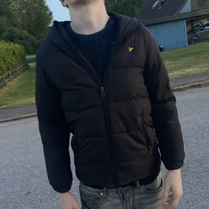 Svart pufferjacka från Lyle & Scott storlek S  - Svart pufferjacka med huva från Lyle & Scott. Jackan har gul logga på bröstet, dragkedja framtill och två sidofickor med dragkedja. Klassisk quiltad look och långärmad modell, perfekt för kyligare dagar.