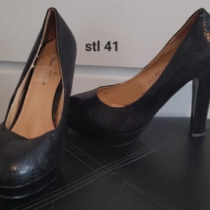 Svarta pumps med platå och klack - Snygga svarta pumps med hög klack och platåsula. Skorna har rund tå och ett diskret ormskinnsmönster som ger en cool touch. Perfekta för dig som vill sticka ut lite extra.