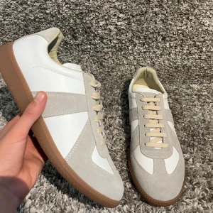 Maison margiela gats - Stilrena sneakers i vitt och beige med ovandel i skinn och mocka, samt klassisk brun gummisula. Skorna har rund tå, platt sula och snörning framtill. Perfekta för dig som gillar minimalistisk och clean stil.