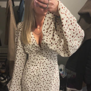 Vit långklänning med körsbär från Adoore - Supersöt vit kortklänning från Adoore med röda körsbärsmönster, v-ringning och markerad midja. Klänningen har långa puffärmar och knappar framtill. Perfekt för sommarens alla tillfällen och ger en riktigt fräsch vibe.