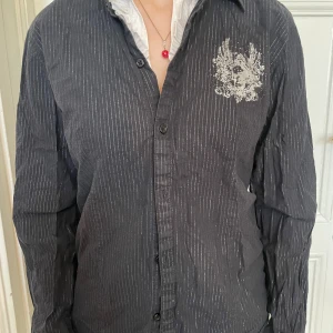 Svart randig skjorta med tryck - Svart skjorta med tunna vita ränder och ett coolt vitt tryck med döskalle och vingar på bröstet. Klassisk krage, knäppning framtill och lång ärm. Skjortan har en lite loose passform och är perfekt att styla lager på lager. Det hade inte skadat att stryka den…