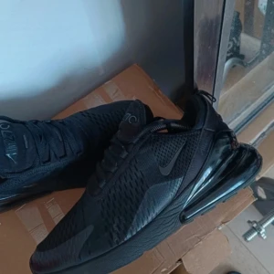 Svarta Nike Air Max 270 sneakers - Säljer ett par helsvarta Nike Air Max 270 sneakers med mesh-ovandel och stor synlig Air-enhet i hälen. Skorna har diskret Nike-logga på sidan och AIR 270-detaljer på plösen och hälen. Perfekta för dig som gillar sportig och clean stil.