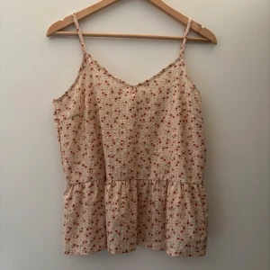 Blommig beige linnetopp med volang - Supersöt topp  med små röda blommor och tunna axelband. Toppen har en volangkant nedtill som ger en luftig och romantisk känsla. Perfekt för varma dagar och enkel att matcha med jeans eller kjol.
