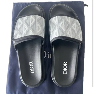 Christian Dior Slides – Dior Atlas Oblique Grå - Säljer ett par exklusiva Dior-sandaler i svart med grå Dior Oblique-tygdetalj. Modellen är stilren och lyxig, perfekt till både vardag och sommarens mer uppklädda tillfällen.  ✔️ Levereras med: 	•	Original Dior-box 	•	Dustbag (skyddspåse) 	•	Äkthetskort 	•	Kvitto & inköpsdokumentation  📏 Storlek: 41 (passar 40-42)      Skick: Ny  💯 Garanterat äkta – komplett med allt som medföljer. 