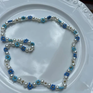 Pärlhalsband med blå blommor - Supergulligt halsband med vita pärlor och små blommor i blått, turkost och vitt. Handgjort! Bandet är elastiskt vilket gör att det passar de flesta. Vid frågor är det bara att skriva!
