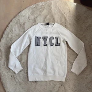 sweatshirt  - Vit sweatshirt med stora grå bokstäver 'NYCL' framtill. Tröjan har rund halsringning, långa ärmar och ribbade muddar. Tillverkad i mjuk bomull, perfekt för en chill och avslappnad stil. En liten fläck på sista bilden