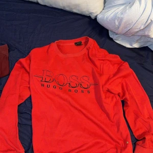 Röd långärmad tröja från Hugo Boss - Snygg röd långärmad tröja från Hugo Boss med stort svart BOSS-tryck på bröstet. Klassisk rund halsringning och mjukt material som känns skönt mot huden. Perfekt för dig som gillar stilrena loggoplagg.