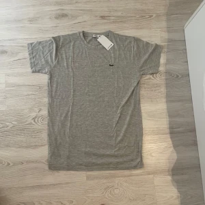 Grå t-shirt från Lacoste - Snygg grå t-shirt från Lacoste med klassisk krokodillogga broderad på bröstet. T-shirten har rund halsringning och korta ärmar. Tillverkad i mjuk bomull som känns skön mot huden. Perfekt för en clean och stilren look.