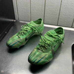 Gröna Nike fotbollsskor strl 41 - Säljer ett par gröna Nike fotbollsskor med snörning och svart swoosh. Skorna har mönstrad ovandel i syntetmaterial och tunna blå detaljer. Perfekta för dig som vill sticka ut på planen. Lätta och smidiga med låg profil. Köpte för 2900 på unisport, hål i vänstra hälen men det märks inte av när man spelar