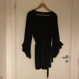 Svart omlott playsuit med volangärm - Supersnygg svart playsuit med omlott-topp, långa ärmar med volangdetaljer och knytband i midjan. Perfekt för dig som gillar en stilren look med lite extra edge. Lätt och flowy känsla, passar till många tillfällen. Bekväm resor i midjan. Superskön. Använd ett fåtal gånger.