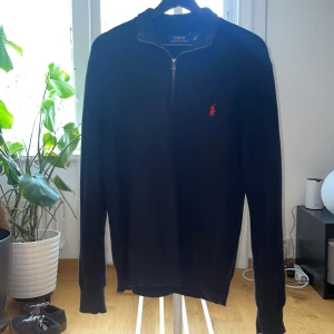 Svart half zip tröja Polo Ralph Lauren - Svart långärmad tröja från Polo Ralph Lauren med röd broderad logga på bröstet. Tröjan har en snygg half zip-dragkedja vid halsen och är tillverkad i ett mjukt stickat material. Perfekt för dig som gillar klassisk och stilren look.