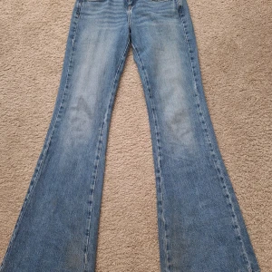 Blå bootcut jeans från Zara, strl 34 - Bootcut jeans från Zara i ljusblå denim med slitna detaljer och råa kanter. Klassisk femficksmodell med hög midja och vida ben. Perfekta för dig som gillar retrovibbar och statement-jeans.