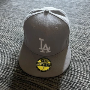 Grå LA New Era 59FIFTY keps - Snygg grå New Era 59FIFTY keps med LA-logga broderad framtill i vitt. Klassisk flat peak och strukturerad passform. Kepsen är helfärgad och har broderad MLB-logga baktill. Perfekt för dig som gillar streetstyle och sportig look.